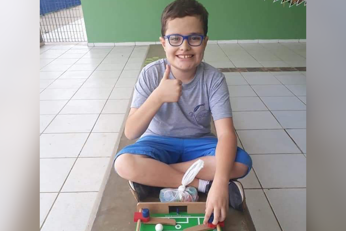 Nicolas, um pré-adolescente com transtorno autista posa para a câmera, sentado no chão, sorrindo e fazendo um joinha.