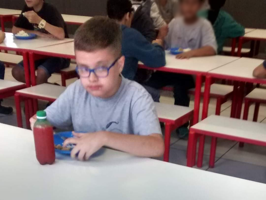 O menino Nicolas, que tem transtorno autista, está sentado sozinho numa mesa de refeitório escolar